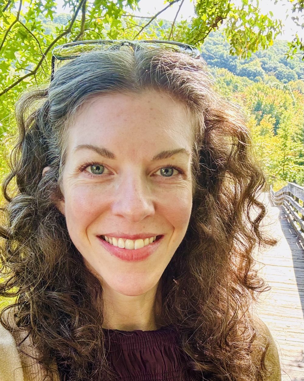 Evelyn Hale smiling in warm sunlight on a wooded boardwalk, green hills behind her