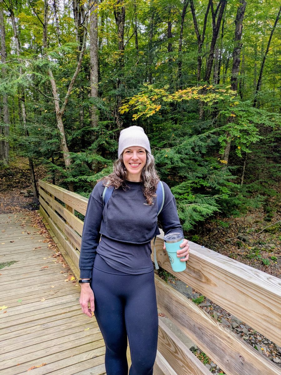 Evelyn in a knit hat on a wooden footbridge in the forest, smiling with a travel mug in hand